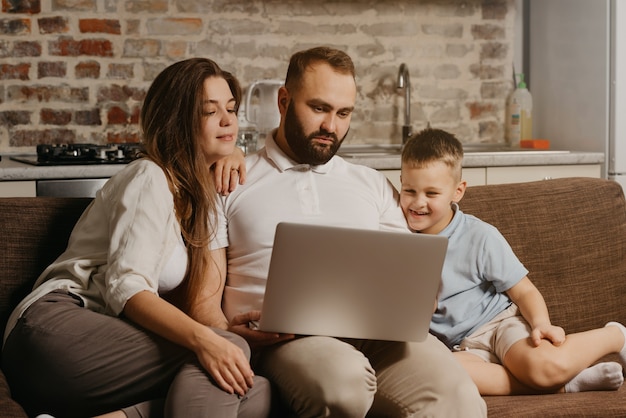 Un padre con barba trabaja de forma remota en una computadora portátil mientras su feliz hijo y su esposa miran la pantalla