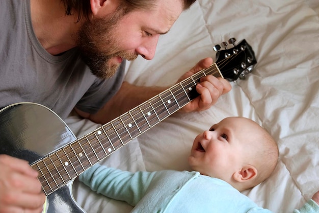 Padre barbudo tocando la guitarra a su hijo pequeño Bebé mirándolo escuchando y sonriendo Retrato de familia feliz en casa Ocio alegre Guapo padre caucásico y lindo bebé divirtiéndose