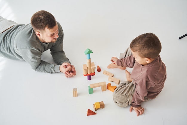Padre cariñoso ayuda a su hijo a jugar en el suelo sobre fondo blanco Padre e hijo construyen una torre de coloridos ladrillos de madera y se divierten juntos