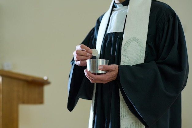 Padre da igreja católica em batina segurando uma xícara pequena com pão ázimo