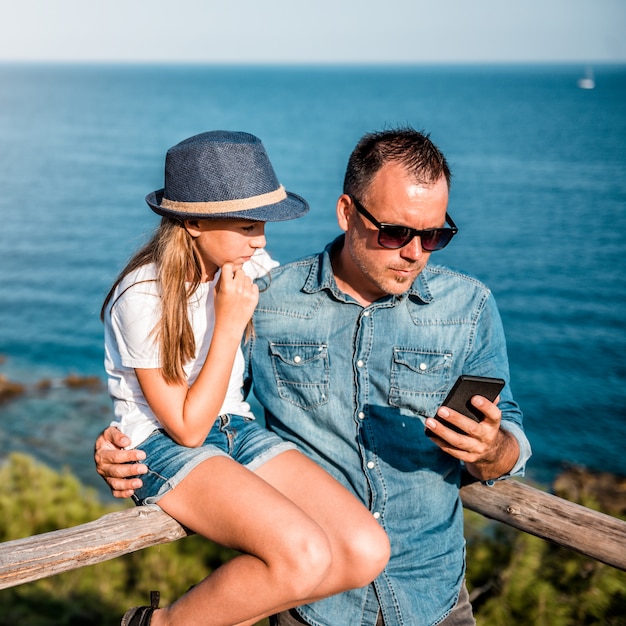 Padre e hija se apoyan contra la valla de madera junto al mar usando un teléfono inteligente