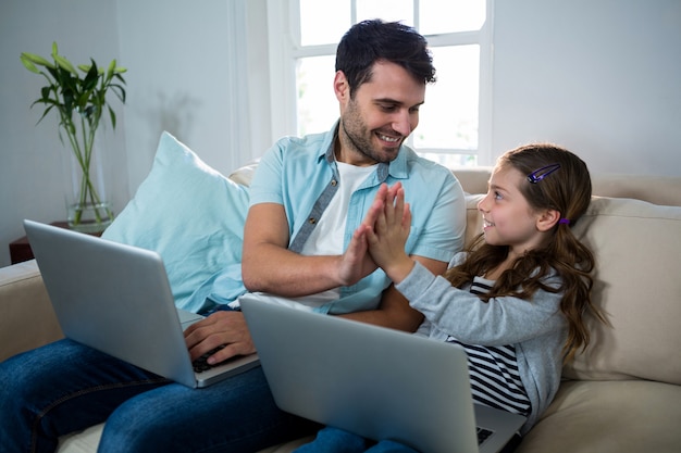 Padre e hija dando cinco mientras usan la computadora portátil en la sala de estar