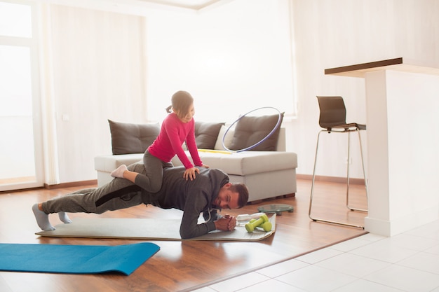 Padre e hija están entrenando en casa. Entrenamiento en el departamento. Deportes en casa. Hacen la tabla de hacer. hija se subió a papá. Él está empujando hacia arriba desde el piso.