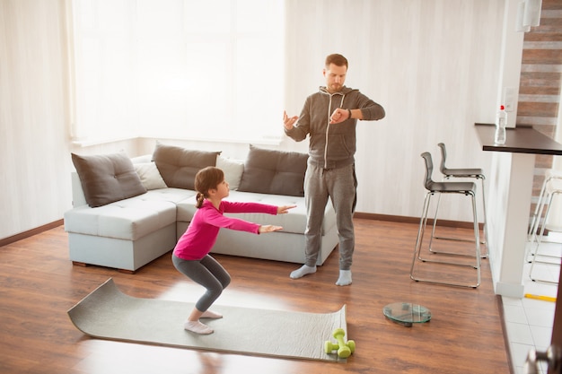 Padre e hija están entrenando en casa. Entrenamiento en el departamento. Deportes en casa. Papá usa un reloj deportivo y su hija está haciendo ejercicios de sentadillas