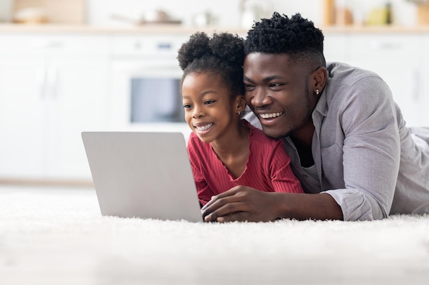 Padre e hija negros alegres que usan la computadora portátil juntos en casa