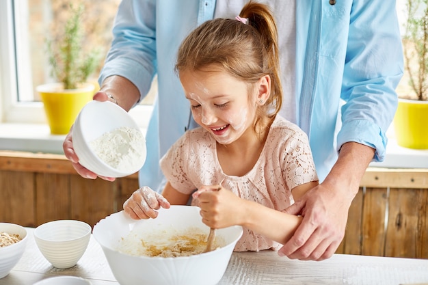 Padre e hija preparando masa juntos en la cocina, agregando harina al cuenco para cocinar pasteles en casa