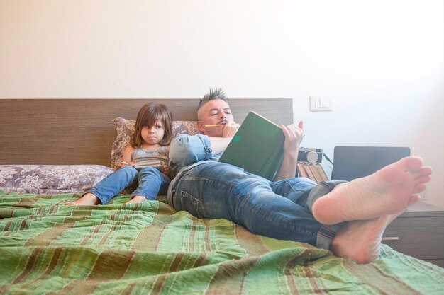 Padre e hija con ropa informal de mezclilla están sentados juntos en la cama la niña está molesta y tal vez aburrida mientras el padre intenta leer un libro concepto de infancia y paternidad