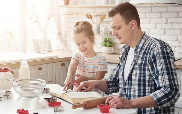 Padre e hija sonrientes leyendo la receta en el libro de cocina en la cocina. Pasatiempo familiar, tarta de cocina u otra panadería, espacio de copia