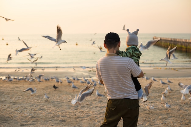 Padre e hijo alimentan gaviotas en la playa al atardecer en Dubai. Hombre con niño alimentar pájaros en la playa