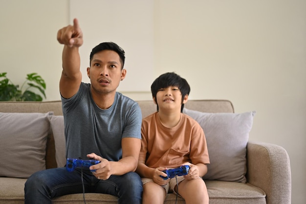 Padre e hijo asiáticos alegres con joysticks están jugando videojuegos juntos en casa