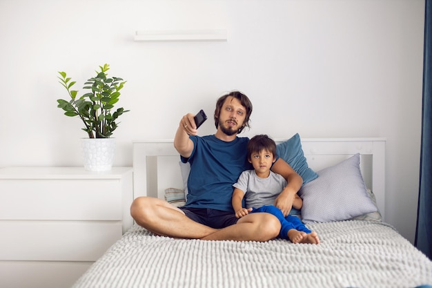 Padre e hijo barbudos están sentados en la cama con camisetas y viendo un partido de fútbol en la televisión. un hombre sostiene un control remoto en sus manos para cambiar de canal