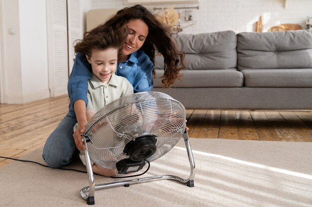 Padre e hijo en casa alegre joven madre con hijo pequeño disfrutan soplando un ventilador de viento fresco en el interior