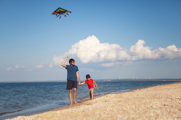 Padre e hijo están de pie en una playa de arena junto al mar y lanzan una cometa rayada de juguete en el verano de vacaciones