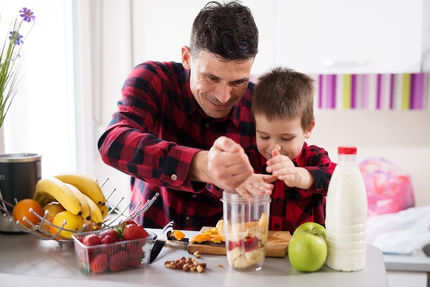 Foto padre e hijo con fuerzas conjuntas están llenando el vaso de la licuadora con frutas en la brillante cocina.