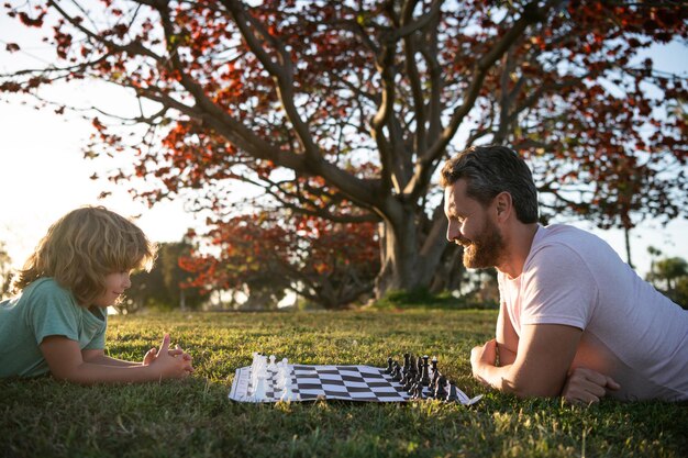 Padre e hijo jugando al ajedrez en el césped en el parque. dia del padre. familia feliz. paternidad e infancia. mate. pasando tiempo juntos. estratégico y táctico. tutoría. papá y el niño juegan al juego de lógica.