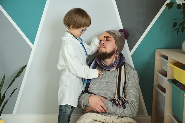 Foto padre e hijo jugando clínica y doctor niño pequeño en bata médica con estetoscopio trata a papá