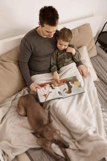 Padre e hijo leyendo un libro en la cama en casa. Hombre atractivo joven y niño con perro descansando en el dormitorio. Colores naturales de la tierra. Ambiente acogedor. El padre lee un cuento de hadas para su hijo.