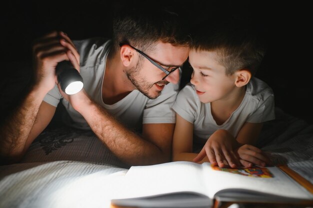 Padre e hijo con linterna leyendo un libro bajo una manta en casa