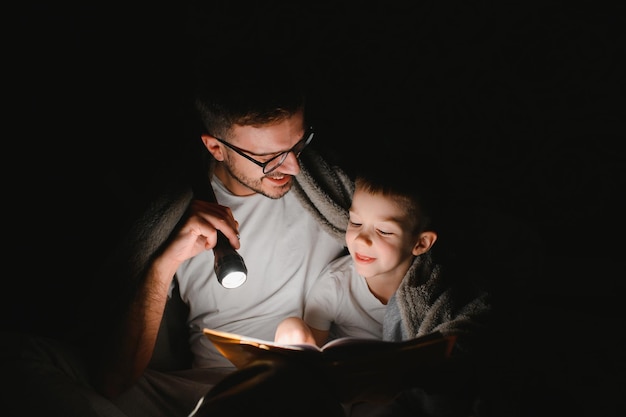 Padre e hijo con linterna leyendo un libro bajo una manta en casa