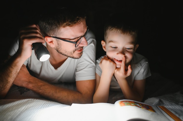 Padre e hijo con linterna leyendo un libro bajo una manta en casa