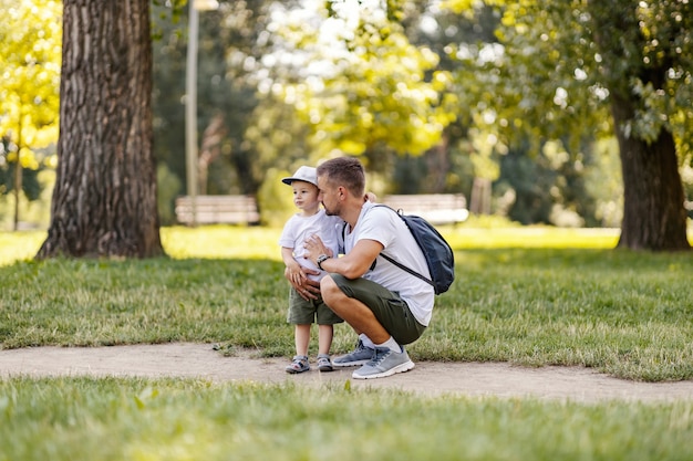 Padre e hijo en la naturaleza. Papá se pone en cuclillas junto al niño y le dice algo mientras están en el bosque en un día soleado. Vestidos con la misma ropa informal, pasan el fin de semana juntos en el parque.