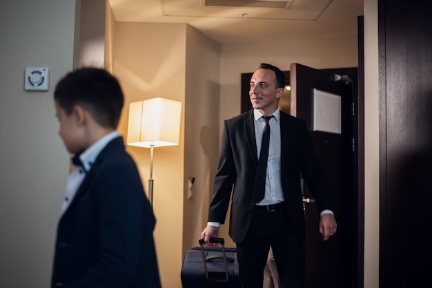 Padre e hijo con ropa formal entrando a una habitación de hotel, el padre lleva una maleta grande