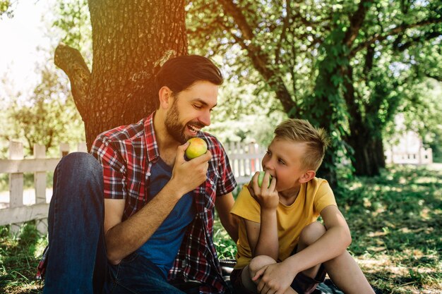 Foto padre e hijo sentados juntos bajo un árbol y comiendo manzana