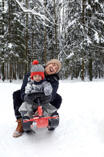 Padre e hijo con sombrero divertido divirtiéndose y riendo en el bosque nevado de invierno Familia feliz vistiendo ropa de abrigo de invierno disfrutando del invierno en un bosque de pinos cubierto de nieve Actividades al aire libre con niños