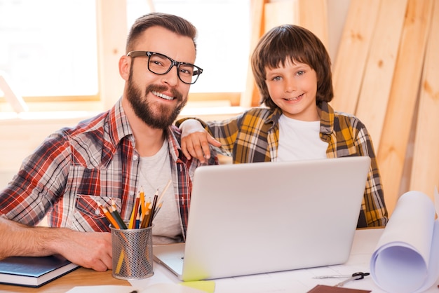 Padre e hijo trabajando juntos. Hombre joven alegre que trabaja en la computadora portátil con su hijo mientras está sentado en el lugar de trabajo