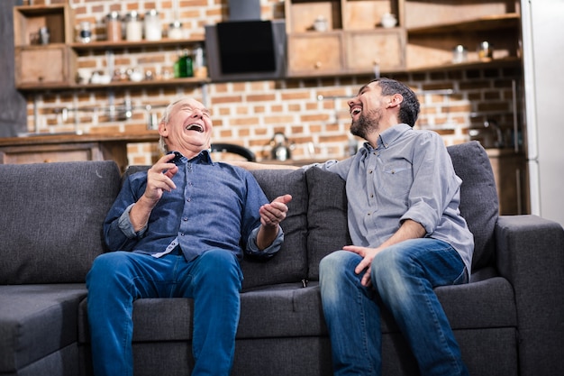 Padre de edad alegre y su hijo haciendo bromas mientras está sentado en el sofá