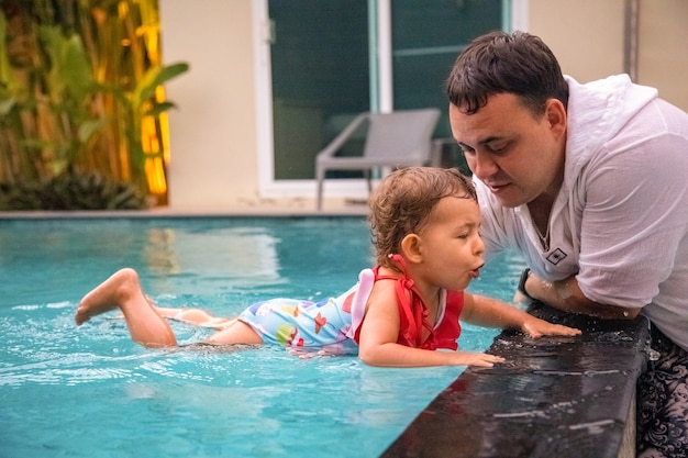 padre enseña a una linda niña pequeña en traje de baño a nadar en la piscina sosteniendo su mano.