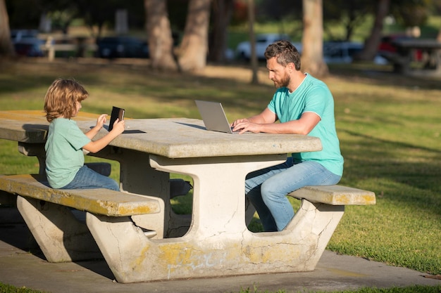 Padre enseñando a su hijo a usar una computadora portátil papá y un niño escolar mirando la pantalla de la computadora y la tableta jugando viendo videos sentados en el césped Fin de semana familiar al aire libre