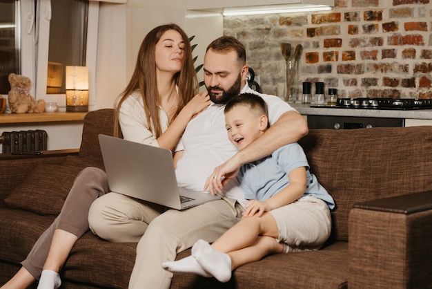 Un padre está tratando de trabajar de forma remota en una computadora portátil cerca de su hijo sonriente y una esposa curiosa en casa