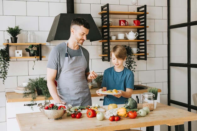 padre de familia, joven e hijo, adolescente, cocinan ensalada de verduras en la cocina y pasan tiempo de calidad juntos, padre e hijo hablando y cocinando comida vegetariana y haciendo tareas domésticas el 8 de marzo y el día de la madre