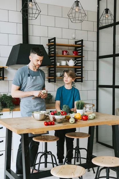 padre de familia, joven e hijo, adolescente, cocinan ensalada de verduras en la cocina y pasan tiempo de calidad juntos, padre e hijo hablando y cocinando comida vegetariana y haciendo tareas domésticas el 8 de marzo y el día de la madre