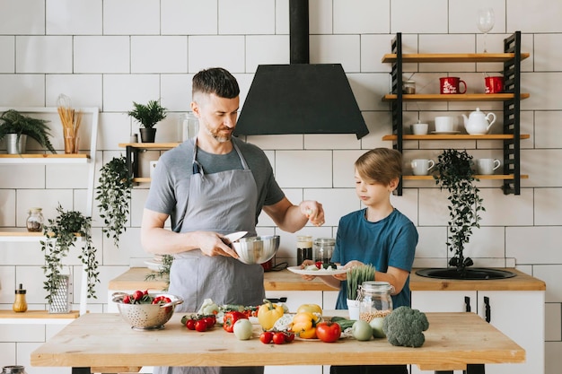 padre de familia, joven e hijo, adolescente, cocinan ensalada de verduras en la cocina y pasan tiempo de calidad juntos, padre e hijo hablando y cocinando comida vegetariana y haciendo tareas domésticas el 8 de marzo y el día de la madre