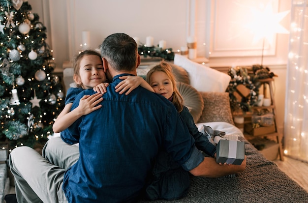 un padre feliz abraza a dos hijas pequeñas sentadas en una cama cerca de un árbol de Navidad felicidad familiar