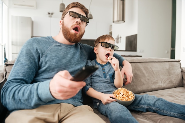Padre feliz confundido sosteniendo el control remoto mientras ve la televisión con su pequeño y lindo hijo usando gafas 3d con palomitas de maíz.