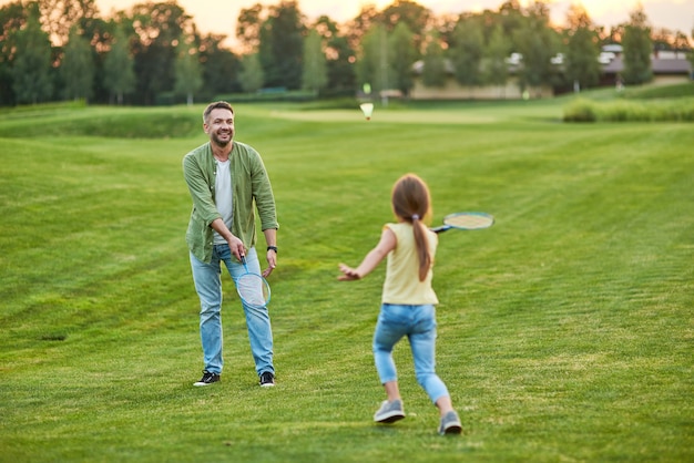 Padre feliz jugando al bádminton con su pequeña hija alegre al aire libre en el parque en un día de verano