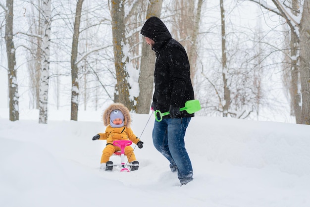 Un padre feliz tira de un gato de nieve para niños a través de la nieve con un niño alegre sentado en él con ropa cálida amarilla