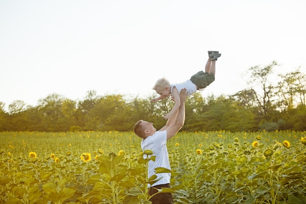 Padre feliz vomitando a un pequeño hijo riendo en un campo verde de girasoles.