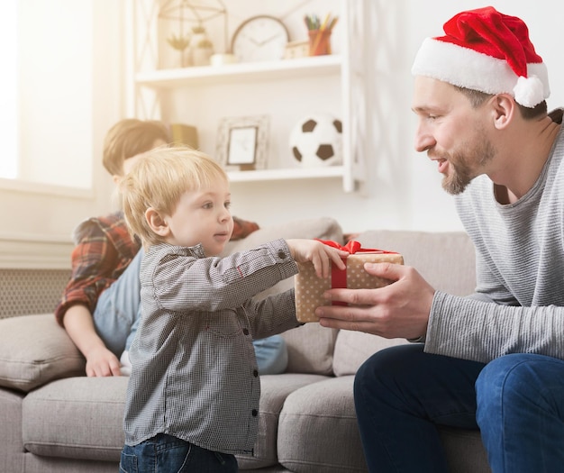 Padre con gorro de Papá Noel dando a su pequeño hijo un regalo de Navidad en casa en la sala de estar