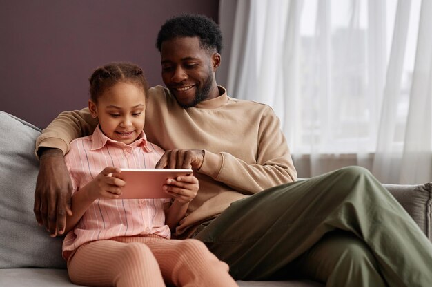 Padre y hija negros usando tableta digital sentados en el sofá en casa y