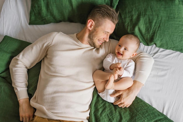 Un padre joven y apuesto se acuesta en la cama en el dormitorio y abraza a su amoroso niño de seis meses que se ríe