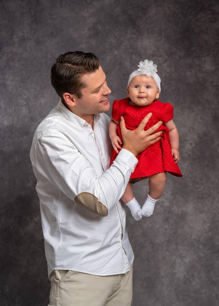 Un padre joven con una camisa ligera sostiene suavemente a su pequeña hija vestida con un vestido rojo en sus brazos Studio disparando el amor del padre por el bebé recién nacido