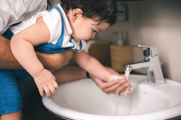 Padre joven y guapo levantando a la pequeña hija mientras la capacita sobre la limpieza ayudándola a lavarse las manos con agua corriente en el lavabo del baño