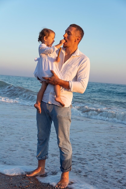 Un padre joven con una hija pequeña camina por la playa al atardecer