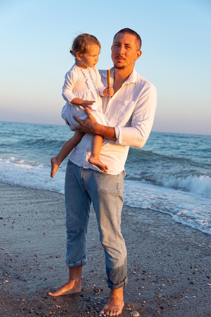 Un padre joven con una hija pequeña camina por la playa al atardecer