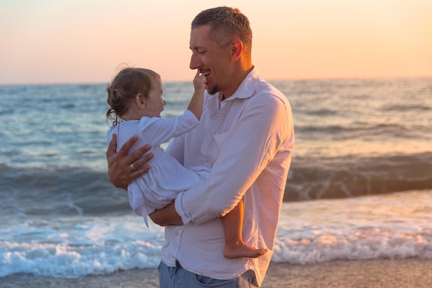 Un padre joven con una hija pequeña camina por la playa al atardecer