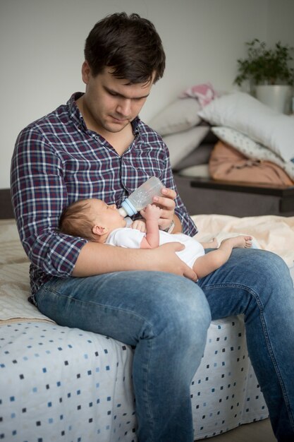 Foto padre joven sentado en la cama y alimentando a su hijo recién nacido